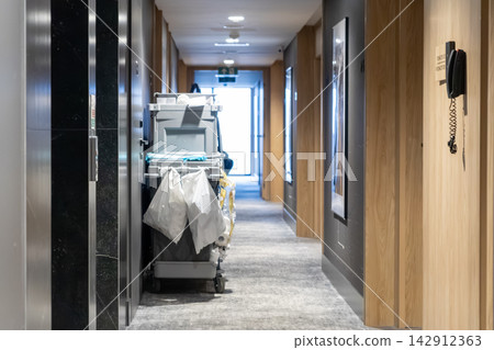 A housekeeping cart stands inside a modern hotel corridor during daily cleaning service, highlighting essential maintenance operations within a professional hospitality facility A housekeeping cart stands inside a modern hotel corridor during daily cleaning service, highlighting essential maintenance operations within a professional hospitality facility 142912363