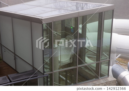 A modern glass water park tower entrance stands outdoors, housing a staircase for a white tube slide in a recreational facility, embodying leisure and adventure for summer vacation fun A modern glass water park tower entrance stands outdoors, housing a staircase for a white tube slide in a recreational facility, embodying leisure and adventure for summer vacation fun 142912366