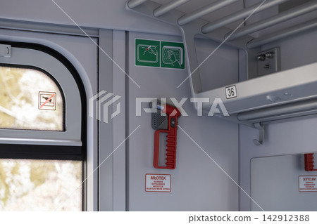 A red emergency glass hammer mounted inside a passenger train carriage wall, illustrating mandatory safety equipment and transit regulation compliance in public rail systems A red emergency glass hammer mounted inside a passenger train carriage wall, illustrating mandatory safety equipment and transit regulation compliance in public rail systems 142912388