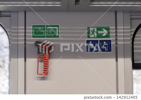 A red emergency hammer is mounted on the interior train wall near safety and accessibility icons, providing vital tools for crisis situations within a modern public transit vehicle A red emergency hammer is mounted on the interior train wall near safety and accessibility icons, providing vital tools for crisis situations within a modern public transit vehicle 142912405