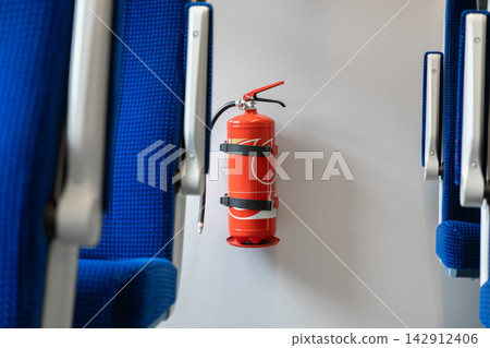 A bright red portable fire extinguisher mounted on a white wall between blue train passenger seats, providing essential emergency protection for commuters within public transport space A bright red portable fire extinguisher mounted on a white wall between blue train passenger seats, providing essential emergency protection for commuters within public transport space 142912406