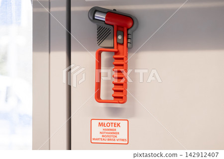 A red emergency glass breaker hammer mounted on a white wall of a modern train carriage, symbolizing passenger safety protocols and essential travel equipment for crisis situations A red emergency glass breaker hammer mounted on a white wall of a modern train carriage, symbolizing passenger safety protocols and essential travel equipment for crisis situations 142912407