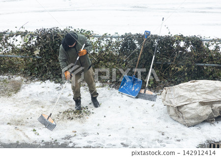 A worker cleaning snow and fallen leaves with a rake on a winter sidewalk, demonstrating professional outdoor maintenance services for public property safety in a cold urban environment A worker cleaning snow and fallen leaves with a rake on a winter sidewalk, demonstrating professional outdoor maintenance services for public property safety in a cold urban environment 142912414