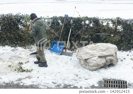 A worker cleans fallen leaves with a rake near a snow-covered hedge and blue plastic shovel, reflecting a cold seasonal routine of park maintenance in a city during late autumn period A worker cleans fallen leaves with a rake near a snow-covered hedge and blue plastic shovel, reflecting a cold seasonal routine of park maintenance in a city during late autumn period 142912415