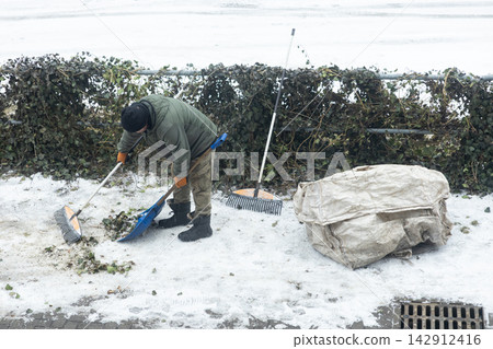 A worker cleaning dead leaves with a rake and shovel on a snowy sidewalk, showcasing diligent municipal landscaping tasks during the cold winter season in an urban city environment 142912416