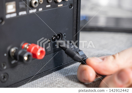 Hand connecting a power cable to an electronic audio device input port, demonstrating technical setup for stereo equipment maintenance within a professional sound production workspace Hand connecting a power cable to an electronic audio device input port, demonstrating technical setup for stereo equipment maintenance within a professional sound production workspace 142912442