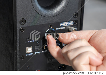 A hand adjusts the volume knob on a black bookshelf speaker panel to control audio levels, highlighting technical setup, sound management, and high fidelity gear in a home studio A hand adjusts the volume knob on a black bookshelf speaker panel to control audio levels, highlighting technical setup, sound management, and high fidelity gear in a home studio 142912447