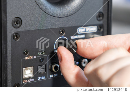 A hand adjusts the volume knob on a professional studio speaker to change audio output levels for a high quality sound experience in a modern music production or home entertainment studio A hand adjusts the volume knob on a professional studio speaker to change audio output levels for a high quality sound experience in a modern music production or home entertainment studio 142912448