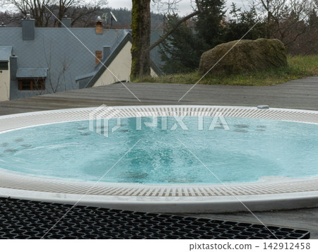 A circular outdoor hot tub sits on a wooden deck by a suburban house, reflecting the peaceful atmosphere of a serene backyard spa retreat designed for relaxation and luxury comfort A circular outdoor hot tub sits on a wooden deck by a suburban house, reflecting the peaceful atmosphere of a serene backyard spa retreat designed for relaxation and luxury comfort 142912458