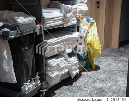 A hotel housekeeping cart sits in a corridor stocked with fresh white linens, folded towels, bathroom amenities, and cleaning supplies for professional room service and maintenance A hotel housekeeping cart sits in a corridor stocked with fresh white linens, folded towels, bathroom amenities, and cleaning supplies for professional room service and maintenance 142912464