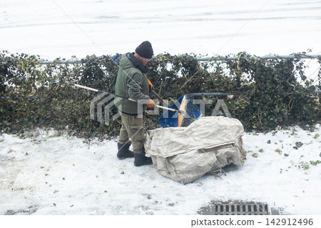 A man in warm winter clothing scoops snow with a blue shovel into a large white fabric sack by a frozen outdoor fence, illustrating maintenance effort in cold icy weather conditions 142912496