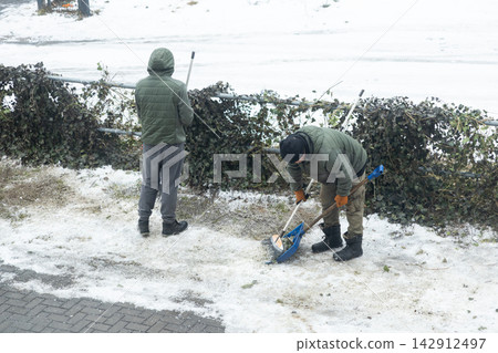 Two workers in warm jackets use a shovel and broom to clear slush and debris from a snowy path, showing community maintenance tasks during cold winter weather in an urban setting Two workers in warm jackets use a shovel and broom to clear slush and debris from a snowy path, showing community maintenance tasks during cold winter weather in an urban setting 142912497