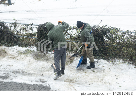 Two workers in green jackets clear snow from a path with shovels near a fence, depicting manual labor and seasonal maintenance during a cold winter day in an urban outdoor setting Two workers in green jackets clear snow from a path with shovels near a fence, depicting manual labor and seasonal maintenance during a cold winter day in an urban outdoor setting 142912498