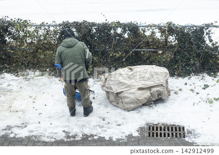 A worker clears snow and debris from a winter street, using a shovel near a white plastic storage bag by a metal fence to ensure public path safety in the cold outdoor environment A worker clears snow and debris from a winter street, using a shovel near a white plastic storage bag by a metal fence to ensure public path safety in the cold outdoor environment 142912499