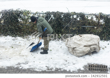 A man in warm clothing clears slush from a path using a blue plastic shovel near a hedge, demonstrating essential winter maintenance and community care during a cold snowy season A man in warm clothing clears slush from a path using a blue plastic shovel near a hedge, demonstrating essential winter maintenance and community care during a cold snowy season 142912501