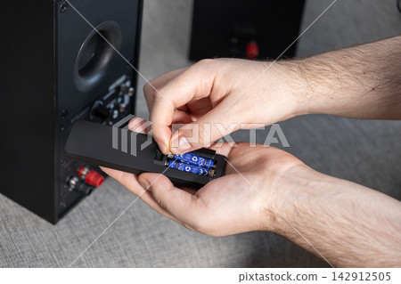 A person installs blue batteries into a black remote control near professional audio speakers, illustrating the maintenance of electronic home entertainment equipment items now A person installs blue batteries into a black remote control near professional audio speakers, illustrating the maintenance of electronic home entertainment equipment items now 142912505