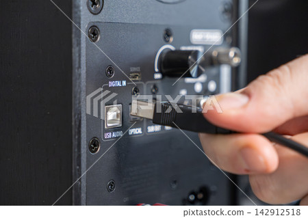 A hand connects a cable to the input port of a modern black speaker, highlighting the technical process of setting up high fidelity home audio equipment for a superior sound system A hand connects a cable to the input port of a modern black speaker, highlighting the technical process of setting up high fidelity home audio equipment for a superior sound system 142912518