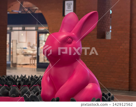 Large bright pink bunny statue stands out among dark animal figurines in a public area, offering a whimsical display that adds playful aesthetic charm to the architectural environment Large bright pink bunny statue stands out among dark animal figurines in a public area, offering a whimsical display that adds playful aesthetic charm to the architectural environment 142912562