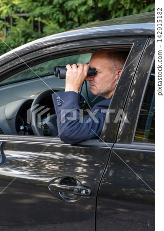 Man in a suit uses binoculars for covert observation of a target from inside a car. Man in a suit uses binoculars for covert observation of a target from inside a car. 142915182