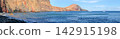Panoramic coastal landscape with rugged red cliffs, blue ocean, anchored sailboats, and dramatic mountains under a cloudy sky, Madeira, Portugal. 142915198