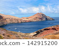 Volcanic coastal landscape with rugged red cliffs, blue ocean, anchored sailboats, and dramatic mountains under a cloudy sky, Madeira, Portugal. 142915200