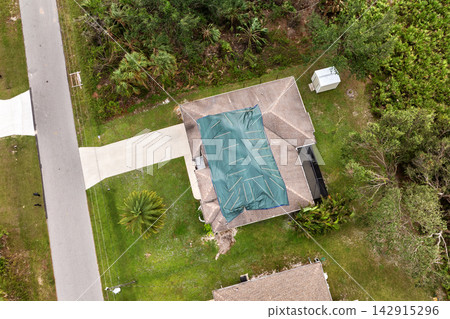 Residential rooftop damaged by hurricane, covered with waterproof tarp sheets to shield interior from rain leaks before roof repairs and shingle replacement are completed Residential rooftop damaged by hurricane, covered with waterproof tarp sheets to shield interior from rain leaks before roof repairs and shingle replacement are completed 142915296