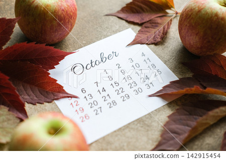 October calendar page on brown background, surrounded by ripe apples and dark red autumn leaves. Harvest season, autumn planning October calendar page on brown background, surrounded by ripe apples and dark red autumn leaves. Harvest season, autumn planning 142915454
