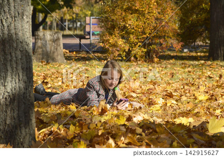Happy girl in a plaid coat lies on yellow leaves and holds a phone in her hands Happy girl in a plaid coat lies on yellow leaves and holds a phone in her hands 142915627