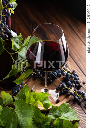 Elegant glass of red wine and bottle surrounded by fresh grapes and leaves on a rustic wooden table 142916042