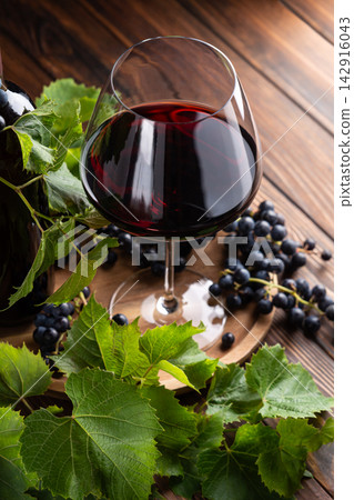 A glass of deep red wine with ripe grapes and fresh green vine leaves on a wooden surface A glass of deep red wine with ripe grapes and fresh green vine leaves on a wooden surface 142916043