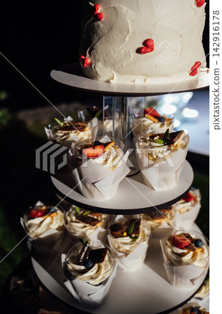 Tiered wedding cake and cupcake tower stand in front of illuminated floral arch at night. 142916178