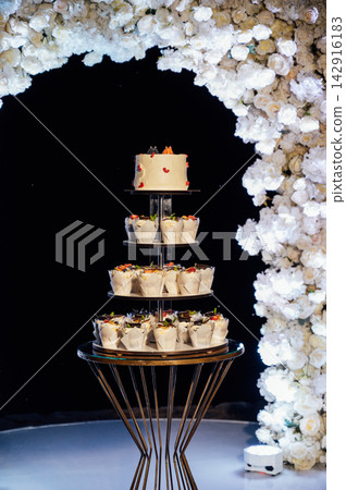 Tiered wedding cake and cupcake tower stand in front of illuminated floral arch at night. Tiered wedding cake and cupcake tower stand in front of illuminated floral arch at night. 142916183