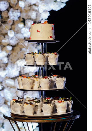Tiered wedding cake and cupcake tower stand in front of illuminated floral arch at night. Tiered wedding cake and cupcake tower stand in front of illuminated floral arch at night. 142916191