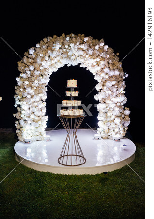 Tiered wedding cake and cupcake tower stand in front of illuminated floral arch at night. Tiered wedding cake and cupcake tower stand in front of illuminated floral arch at night. 142916193