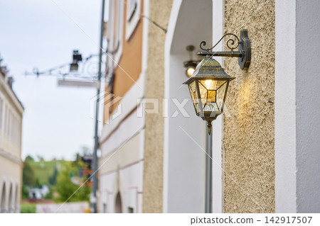 Wall mounted lantern glowing on textured building facade beside narrow street Wall mounted lantern glowing on textured building facade beside narrow street 142917507