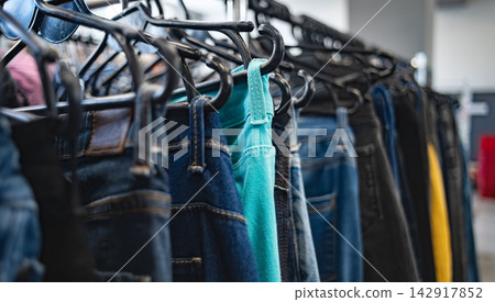 Used denim jeans and colorful tops hanging on metal rack in secondhand clothing store Used denim jeans and colorful tops hanging on metal rack in secondhand clothing store 142917852