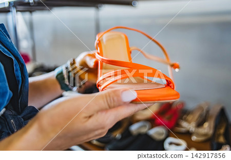 Woman holding bright orange sandal in secondhand shoe rack in thrift shop Woman holding bright orange sandal in secondhand shoe rack in thrift shop 142917856