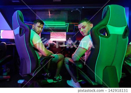 Two Gamers In Neon-Lit Gaming Chairs Competing At A LAN Party With Monitors And Headsets Two Gamers In Neon-Lit Gaming Chairs Competing At A LAN Party With Monitors And Headsets 142918131