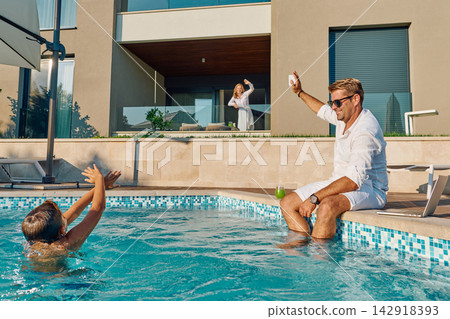 Family Enjoying Luxury Poolside Morning: Man Waving to Woman on Balcony While Child Plays in Water 142918393
