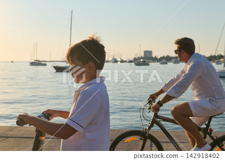 Father And Son Riding Bikes And Scooter Along Marina Waterfront At Sunset Father And Son Riding Bikes And Scooter Along Marina Waterfront At Sunset 142918402