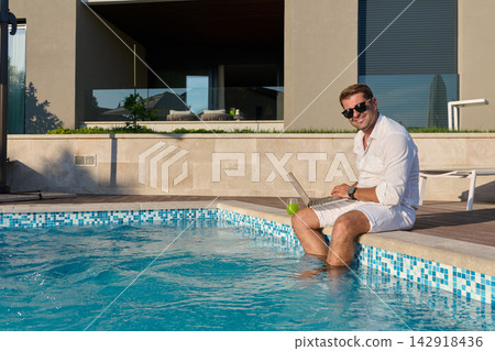 Man Working Remotely by Private Pool on Sunny Summer Morning With Laptop and Drink Man Working Remotely by Private Pool on Sunny Summer Morning With Laptop and Drink 142918436