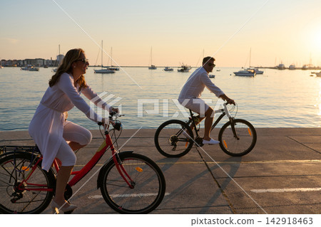 Couple Riding Bicycles Along Seaside Promenade at Sunset Near Marina and Boats 142918463
