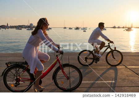 Couple Cycling Along Seaside Promenade At Sunset On Vacation For Relaxed Romantic Outdoor Leisure Couple Cycling Along Seaside Promenade At Sunset On Vacation For Relaxed Romantic Outdoor Leisure 142918469