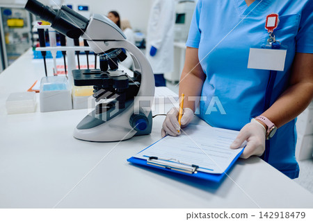 Lab technician preparing a report while using a microscope in a modern laboratory setting Lab technician preparing a report while using a microscope in a modern laboratory setting 142918479