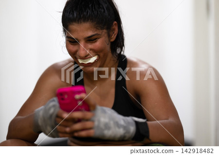 Young Female Boxer Wearing Mouthguard and Hand Wraps Smiling While Checking Phone After Workout Young Female Boxer Wearing Mouthguard and Hand Wraps Smiling While Checking Phone After Workout 142918482