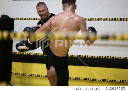 Coach Training Young Fighter With Kicking Pads During Intense Sparring Session In Boxing Ring Coach Training Young Fighter With Kicking Pads During Intense Sparring Session In Boxing Ring 142918565