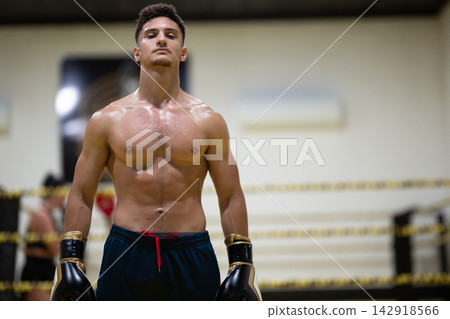 Confident Shirtless Boxer Standing in Boxing Ring With Gloves Ready After Intense Training Session Confident Shirtless Boxer Standing in Boxing Ring With Gloves Ready After Intense Training Session 142918566