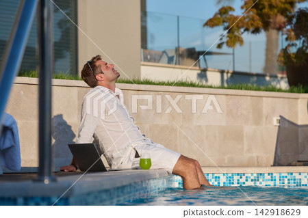 Relaxed Man With Laptop by Poolside Enjoying Sunlight and Green Smoothie While Working Remotely 142918629