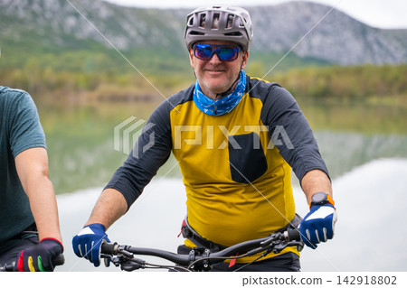 Middle-aged man mountain biking by a lake wearing helmet, sunglasses and yellow jersey Middle-aged man mountain biking by a lake wearing helmet, sunglasses and yellow jersey 142918802