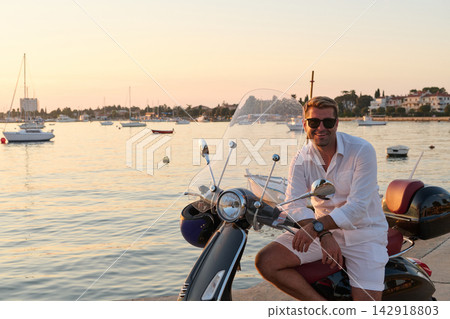 Smiling Man on Scooter by the Seaside at Sunset Enjoying Relaxed Summer Vacation Lifestyle Smiling Man on Scooter by the Seaside at Sunset Enjoying Relaxed Summer Vacation Lifestyle 142918803